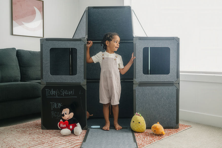 A child playing with the Superspace Big Set, a modular playset made of life-sized magnetic panels in a charcoal color. The playset is built into a fort structure, featuring windows and a chalkboard panel for creative play. Perfect for imaginative adventures, the Big Set encourages open-ended, screen-free fun and is easy to assemble and store.