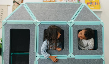 Two children joyfully playing inside a structure built with the Superspace Big Set in its teal variant. The life-sized magnetic panels are configured into a house shape with window openings, encouraging imaginative play and social interaction. Perfect for fostering creativity and collaborative fun.